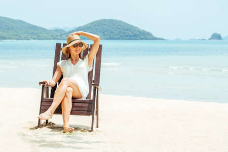 Happy young woman sitting on beach chair on tropical beach in white dress. Enjoying life, carefree,...