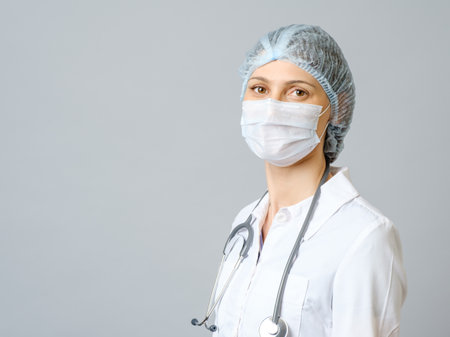 Portrait of young female doctor in protective medical mask on her face and cap on her head. Isolated on gray backgroundの写真素材