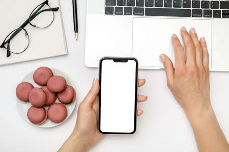 Mock up image of female hand holding and using black mobile phone with blank screen on laptop...