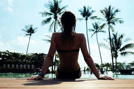 Back view of young woman in swimsuit sitting at a poolside