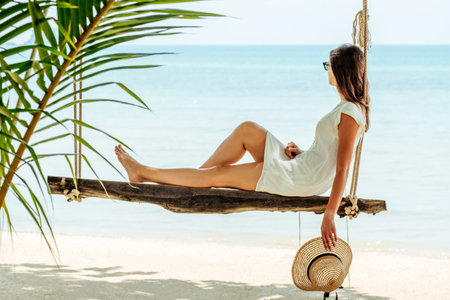 Vacation concept. Young woman in white dress and hat swinging at a beachの写真素材