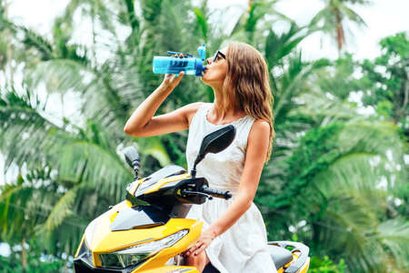 Portrait of young woman on scooter drinking water from reusable bottleの写真素材