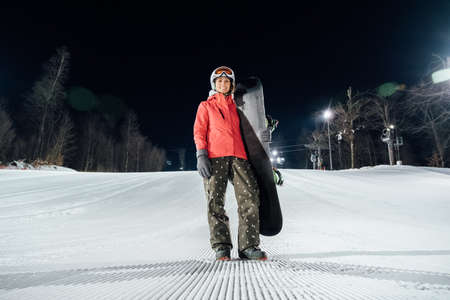 Portrait of female snowboarder wearing white helmet and red jacket standing with snowboard at evening slope. Winter sports conceptの写真素材