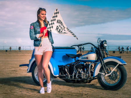Ouistreham, France September 2021 A beautiful girl with a flag in her hand is standing in front of an old motorcycle. Racing on the beach in Normandyのeditorial素材