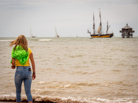 La rochelle, France, July 2022. A young, blonde woman is walking on the beach with a sailing ship and a lighthouse in the background. Relax, vacationのeditorial素材