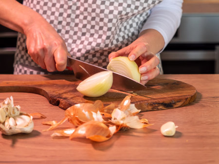 cutting onion on a wooden board, cook in a checked apron with a knife in her hand, wooden table top, front photoの写真素材