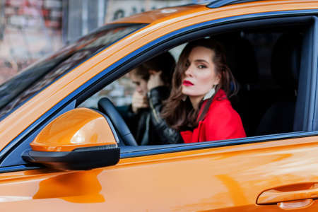 Beautiful girl portrait in a red coat posing in orange car with young man who out of focus. girl in...