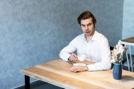 Handsome pensive male in white shirt looking away sitting at table in cafeteria thinking about plans, caucasian guy waiting for meeting in coffee shop dreaming about idea on leisureの写真素材