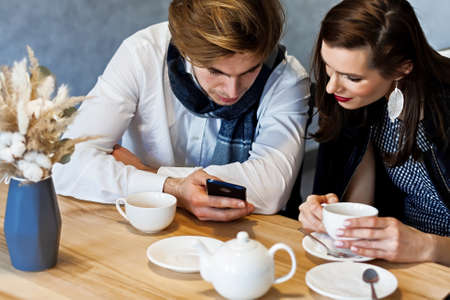 beautiful couple in a cafe at a table drinks coffee and watching something on his phoneの写真素材