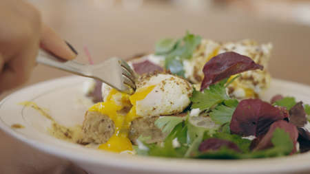 A female hand cutting poached Egg Benedikt so that yellow warm yolk pours out. A delicious salad and egg for breakfast in a restaurant. Healthy meal for anonymous woman. Traditional european brunch. High quality photoの写真素材