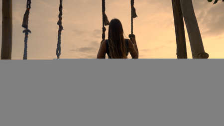 A dark silhouette of a girl on swing at seaside. A beautiful vacation leisure on a beach at sunset near ocean shore. Tropical romance and enjoyable time swinging alone.Anonymous female flying on swingの写真素材
