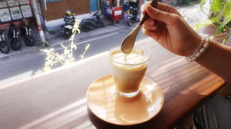 Anonymous girl stirring thick milk foam of a coffee drink on a window sill of a cafe overlooking a Balinese street. Perfect sunny morning for a female on vacation in tropical paradise. Avogato coffee.の写真素材