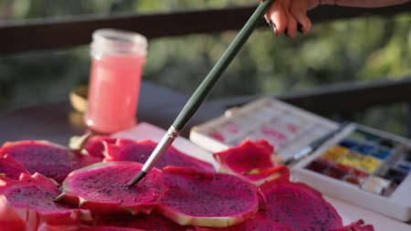Artist painting real tropical fruits with watercolor paint. Funny hyperrealism concept. Contemporary art performance on plein-air with watercolours and easel of sliced dragonfruits in tropical garden.の写真素材
