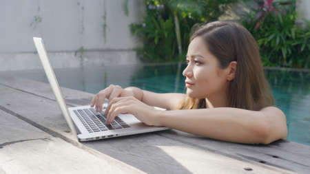 A beautiful mixed-race programmer girl in a blue swimsuit is working on a laptop while standing in the pool. A female freelancer is in the pool typing on a computer keyboard A sunny day in the tropicsの写真素材