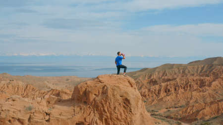 A Caucasian male traveler in a blue T-shirt stands on the top of a mountain and drinks water from a...