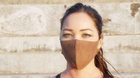 Close-up of a mixed-race girl with beautiful green eyes and a medical mask looking directly into the camera. a serious look of a young girl against the background of a sunny concrete stadium.の写真素材