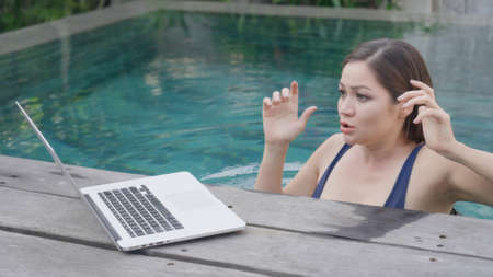 A young mixed-race girl in a blue swimsuit is working at a laptop while standing in the pool. A beautiful girl sees an unpleasant message.の写真素材