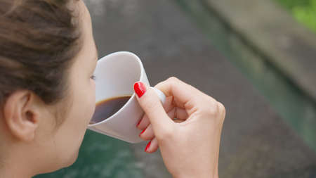 Close-up of a girl with a red manicure drinking coffee from a white cup against the background of a...