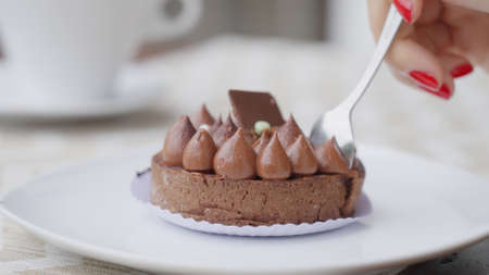 A woman with a beautiful red manicure breaks off a piece of chocolate cake with a teaspoon, a white...
