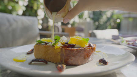 Close-up of an exquisite French toast with orange slices on a white plate. A man pours condensed...