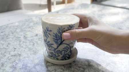 Close-up of a human hand with a pink manicure taking a white coffee mug from a marble restaurant table on a sunny day. The girl takes a white coffee mug with a blue pattern in the style of Gzhel.の写真素材
