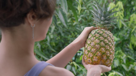 A girl with beautiful earrings jewelry is looking at beautiful pineapple against a background of bright green tropical thickets. A girl with tropical fruit on sunny day. Soft focus Film grain textureの写真素材