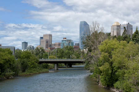 Blue skies reflected in the river with green trees in the foreground to the right and the city highrises towering over the scene.の写真素材