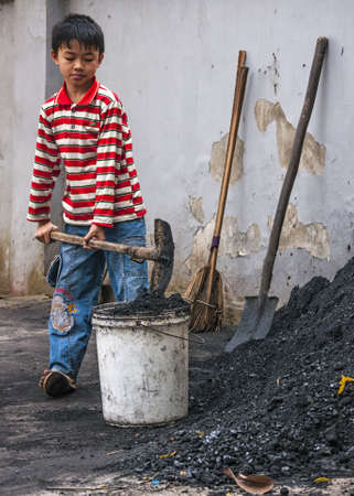 Duong Lam, Vietnam - March 8, 2012: Boy scoops coal in bucket to take inside home. Kid with colorful shirt uses shovel to put black coal in white bucket.のeditorial素材