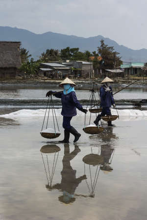 South Vietnam coastline - March 2012: Focus on two masked women carrying baskets of salt and who are reflected in the pool.
