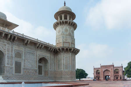 Minaret of Agra s Baby Taj mausoleum with West gate in the background, India  Combination shot of white marble edifice and red white gate building both against blue cloudy skies の写真素材