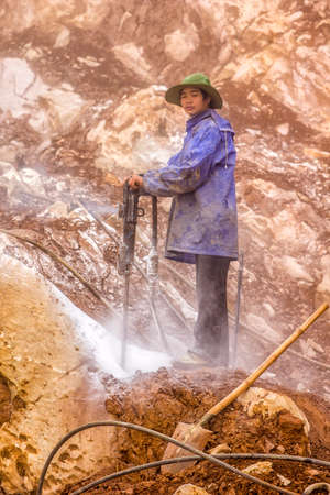 SA PA, VIETNAM - CIRCA MARCH 2012  Roadworker  caught in dust drilling rocks from the cliff  His blue jacket and green hat offset from the brown beige rocks captured in a dusty haze のeditorial素材