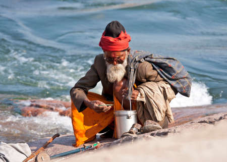 ORCHHA, INDIA - CIRCA FEBRUARY 2011: White bearded old man, a sadhu or holy man, with sunglasses and the classic metal water pot sits on his haunches at the rocky river's edge.のeditorial素材