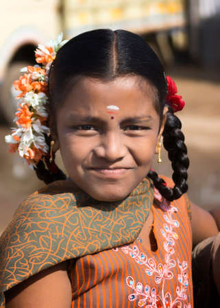KUMBAKONAM, INDIA - OCTOBER 11, 2013: A young Hindu schoolgirl with flowers in her black hair faintly smiles into the camera. In the close-up, her orange shirt and matching scarf accent her face.のeditorial素材