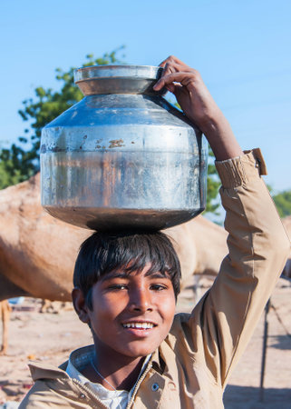 NAGAUR, INDIA - FEBRUARY 10, 2011: A young boy carries a large metal pot of water on  his head. He looks into the camera with a smile.のeditorial素材