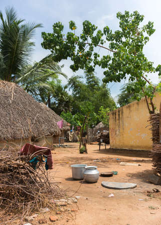 KUMBAKONAM, INDIA - OCTOBER 11, 2013: A group of humble houses, some with straw roofs and mud walls stand at a dusty, sandy walkway. Kitchen utensils are strewn on the ground. Firewood is piled up. Laundry hangs to dry.のeditorial素材