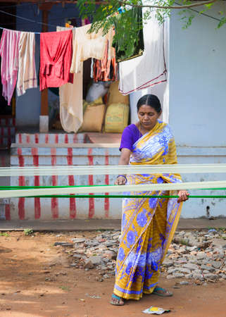 KUMBAKONAM, INDIA - OCTOBER 12, 2013: Middle-aged woman in colorful sari separates drying silk strings that are stretched between two poles. Street scene in front of her house.のeditorial素材