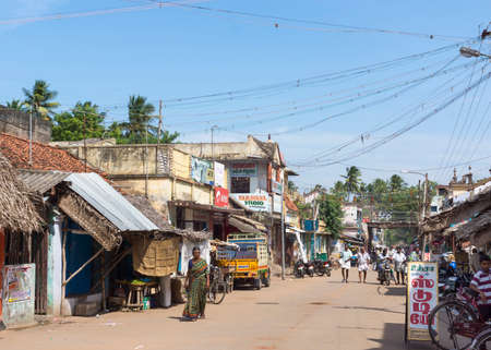 KUMBAKONAM, INDIA - OCTOBER 12, 2013: Street leading to the Mahalingeswarar Temple. Small poor houses with retail activity at the ground floor line the street at both sides. A few people walk up the street. Plenty of electric cables against blue sky.のeditorial素材