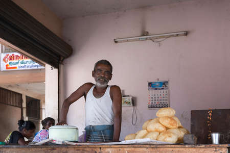 KUMBAKONAM, INDIA - OCTOBER 12, 2013: Smiling Fast food baker with frying pan and a pile of his fried cookies. He stands in his small shop with open windows. Two customers in the photo.のeditorial素材