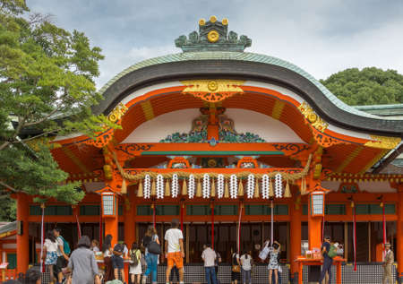Kyoto, Japan - September 17, 2016: People gather in front of the main vermilion shrine at Fushimi Inari Taisha Shinto Shrine. They appeal and pray to the spirits of ancestors. Gray roof with golden trim. Cloudy sky and green trees.のeditorial素材