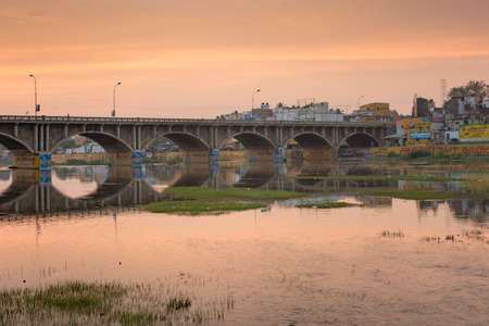 Madurai, India - October 22, 2013: Early morning photo of highway 85 bridge into Madurai over the Vaigai River. Bows of bridge reflected in water. Orange-yellow sky reflected in water. Billboards.のeditorial素材