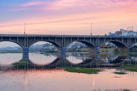 Madurai, India - October 22, 2013: Early morning photo of highway 85 bridge into Madurai over the Vaigai River. Bows of bridge reflected in water. Blue, purple sky reflected, too. Billboards.のeditorial素材