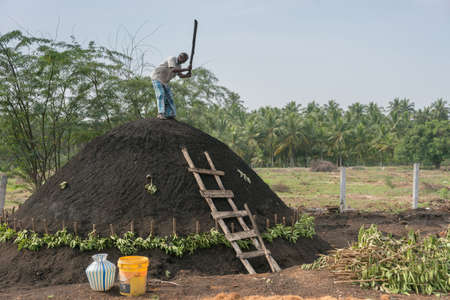 Dindigul, India - October 22, 2013: A man on top of his charcoal hill of black earth which covers raw wood. The man fastens the black dirt by hammering it with a beam. The ancient way. Blue sky and green background.のeditorial素材