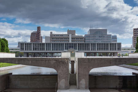 Hiroshima, Japan - September 20, 2016: Seen from behind the pool and the eternal flame stands the long Peace Memorial building and museum. Office buildings tower over it.のeditorial素材