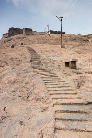 Dindigul, India - October 23, 2013: Stairs hewed in huge boulder leading to historic Dindigul Rock Fort. Brown-beige stone. Fort wall against blue sky.のeditorial素材
