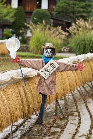 Shirakawago, Japan - September 23, 2016: Closeup of well designed and decorated scarecrow with a neck sign and a butterfly net at Shirakawago. House with particular joined hands roof in the back.のeditorial素材