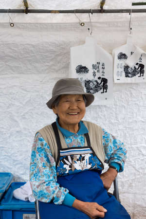 Takayama, Japan - September 24, 2016: Closeup of old smiling lady who is a vendor on the public morning market in front of the Jinya Hall. Blue clothes, white tarp background.のeditorial素材