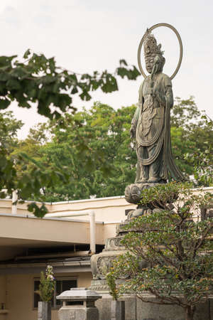 Tokyo, Japan - September 26, 2016: Stone statue of Bodhisattva Quan Yin standing on lotus pedestal in garden at Senso-ji Buddhist Temple. Large halo. Green foliage. Gray sky.のeditorial素材