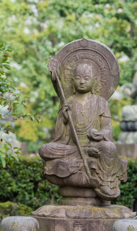 Tokyo, Japan - September 26, 2016: Closeup of stone statue of Bodhisattva sitting on lotus pedestal in garden at Senso-ji Buddhist Temple. Large halo. Holds staff and sphere. Green foliage.のeditorial素材