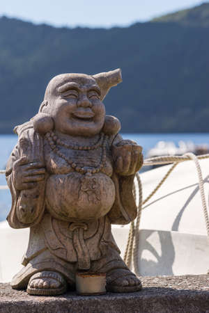Hakone, Japan - September 27, 2016: A wooden statue of the laughing and obese Japanese God Ebisu stands at the shoreline of Lake Ashi. White color and rope of boat, forest and water.のeditorial素材