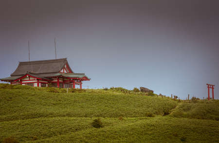 Hakone, Japan - September 27, 2016: Vermilion Mototsumya Shinto shrine on top of green mount kamagatake under dark rain clouds gathering sky. Entrance Torii at other end.のeditorial素材